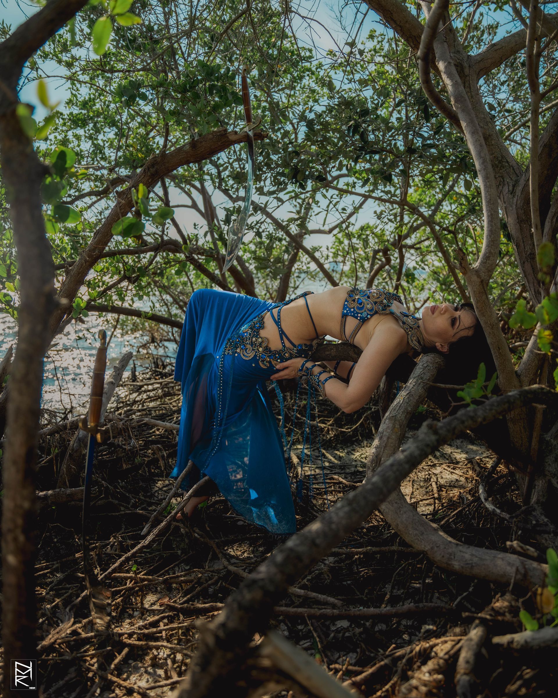 Belly dancer Azeeria Azizah in a blue dress is laying on a tree branch.