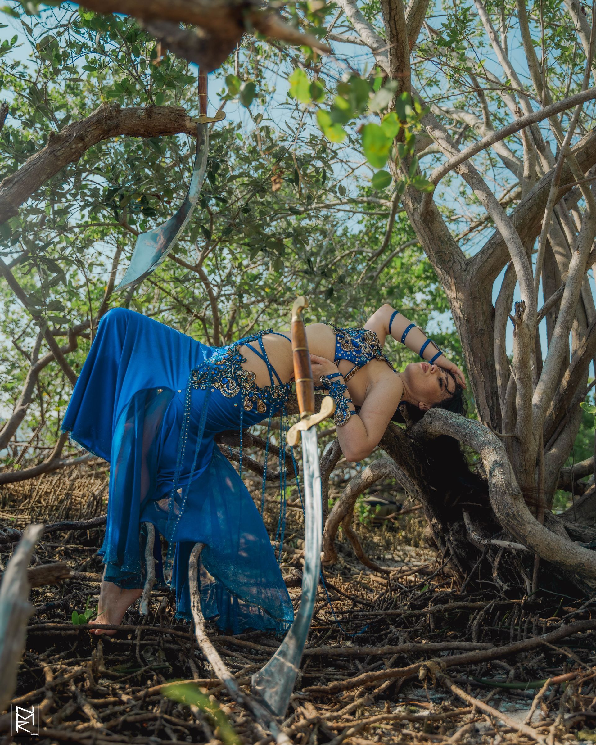 Belly dancer Azeeria Azizah in a blue belly dance costume posing by laying on a tree branch with a sword in front of her.