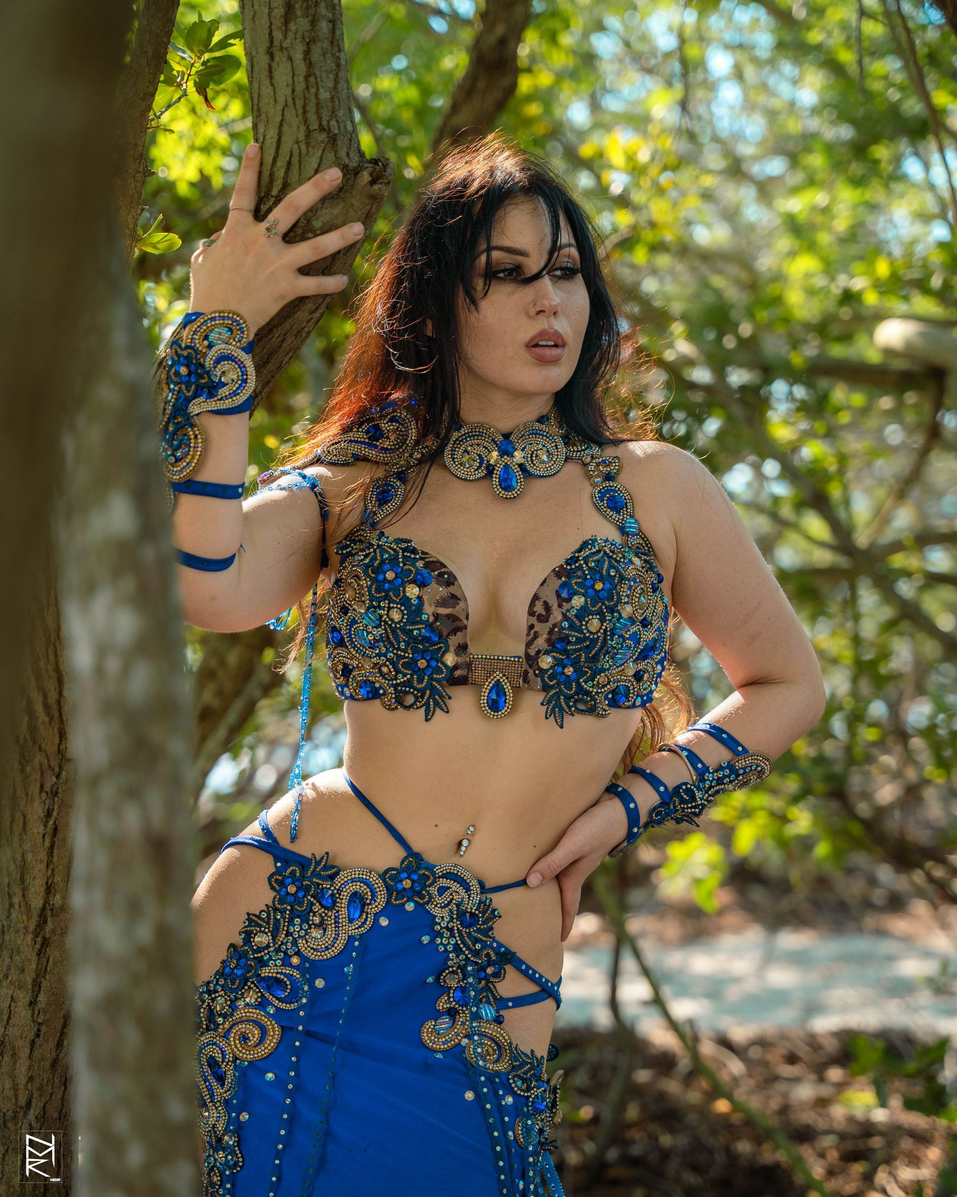 Belly dancer Azeeria Azizah in a blue and gold belly dancer costume standing next to a tree.