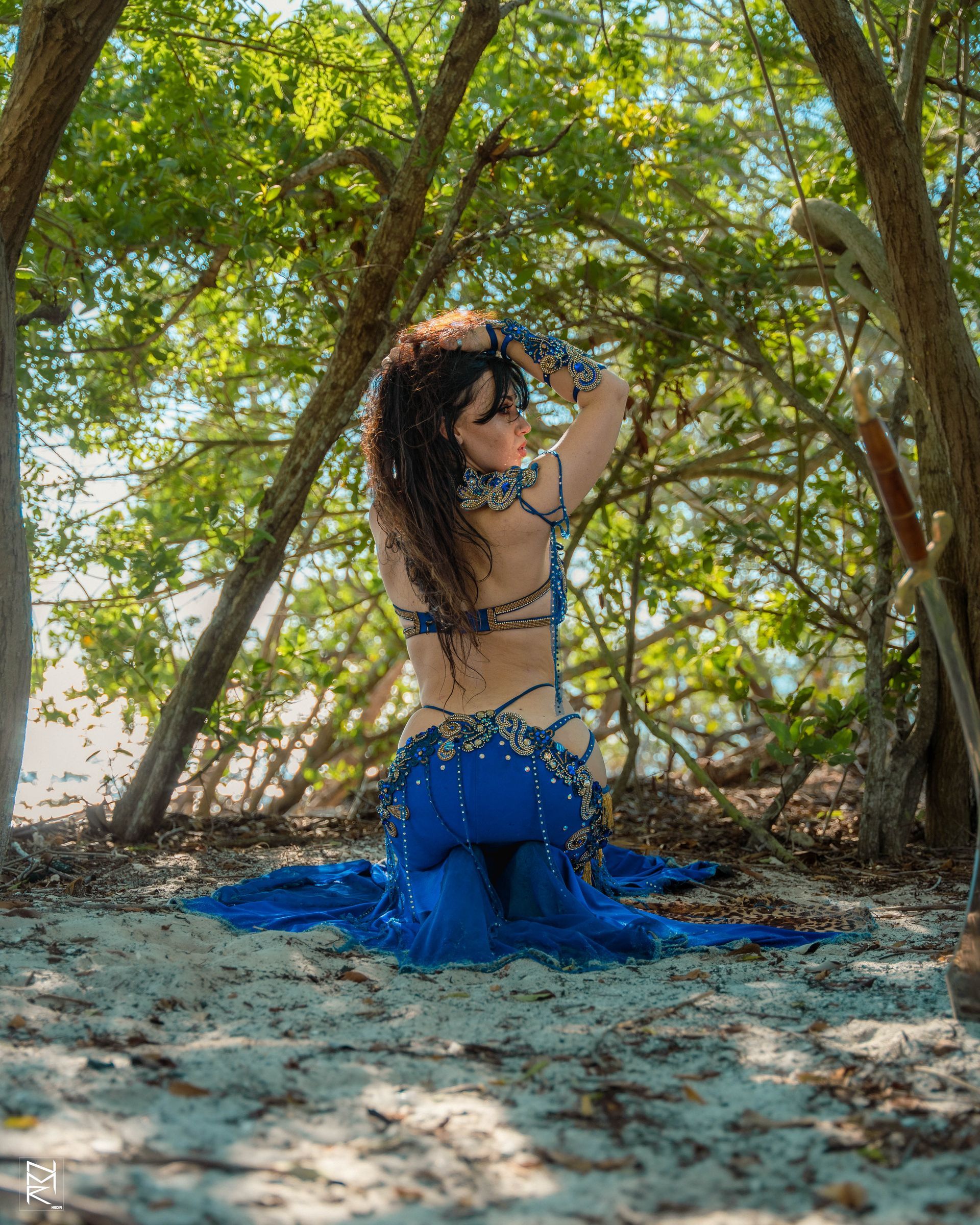 Belly dancer Azeeria Azizah in a blue dress kneeling on the ground in the woods.