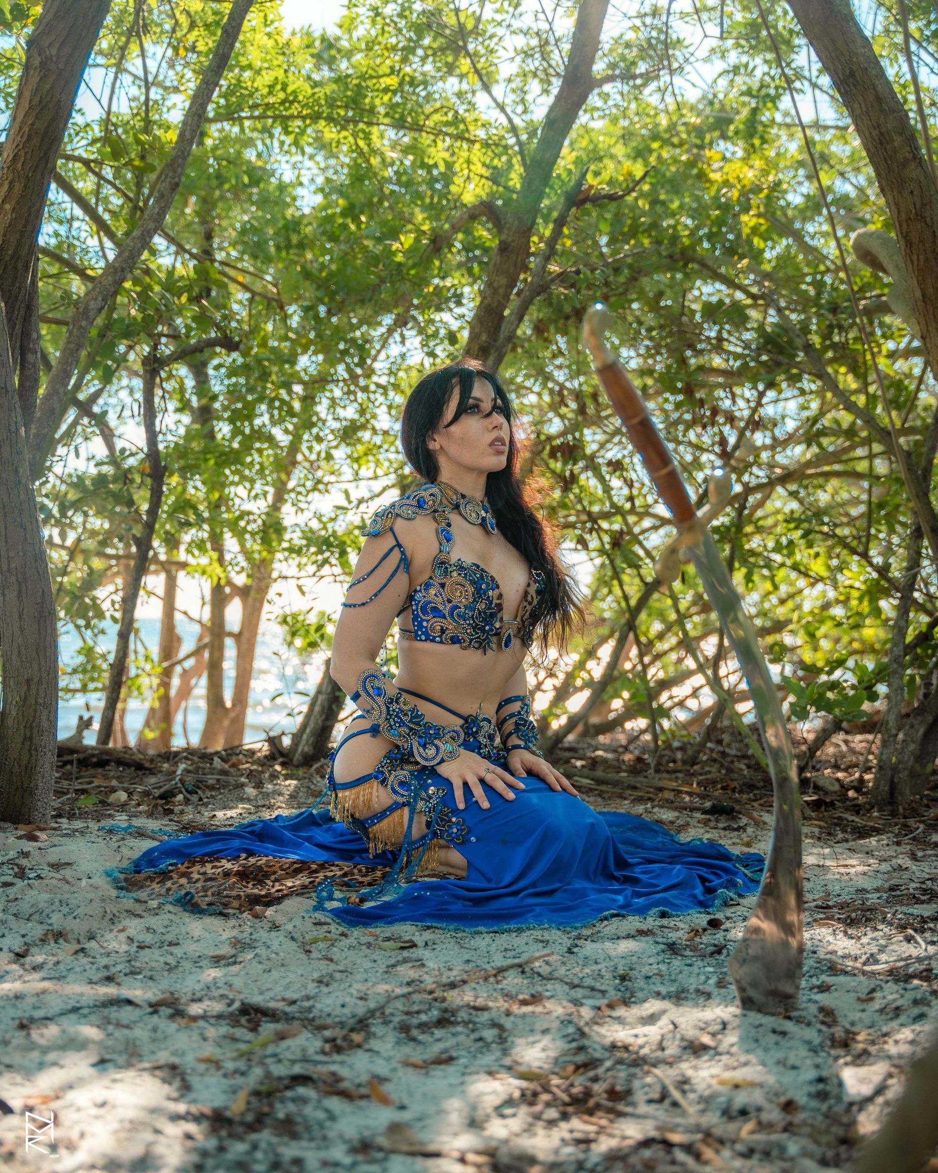 Belly dancer Azeeria Azizah in a blue dress kneeling on the ground in the woods.