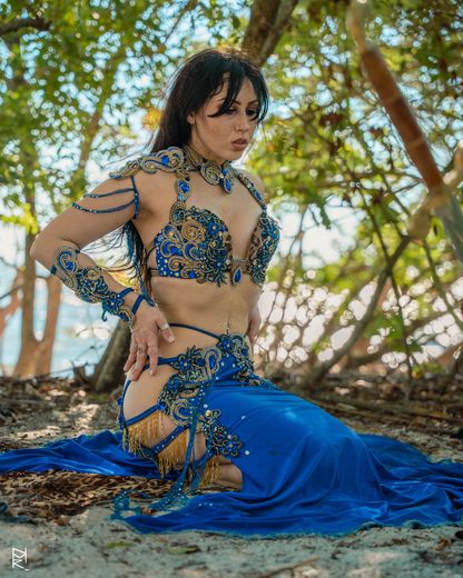 Belly dancer Azeeria Azizah in a blue and gold belly dancer costume is kneeling in the sand.