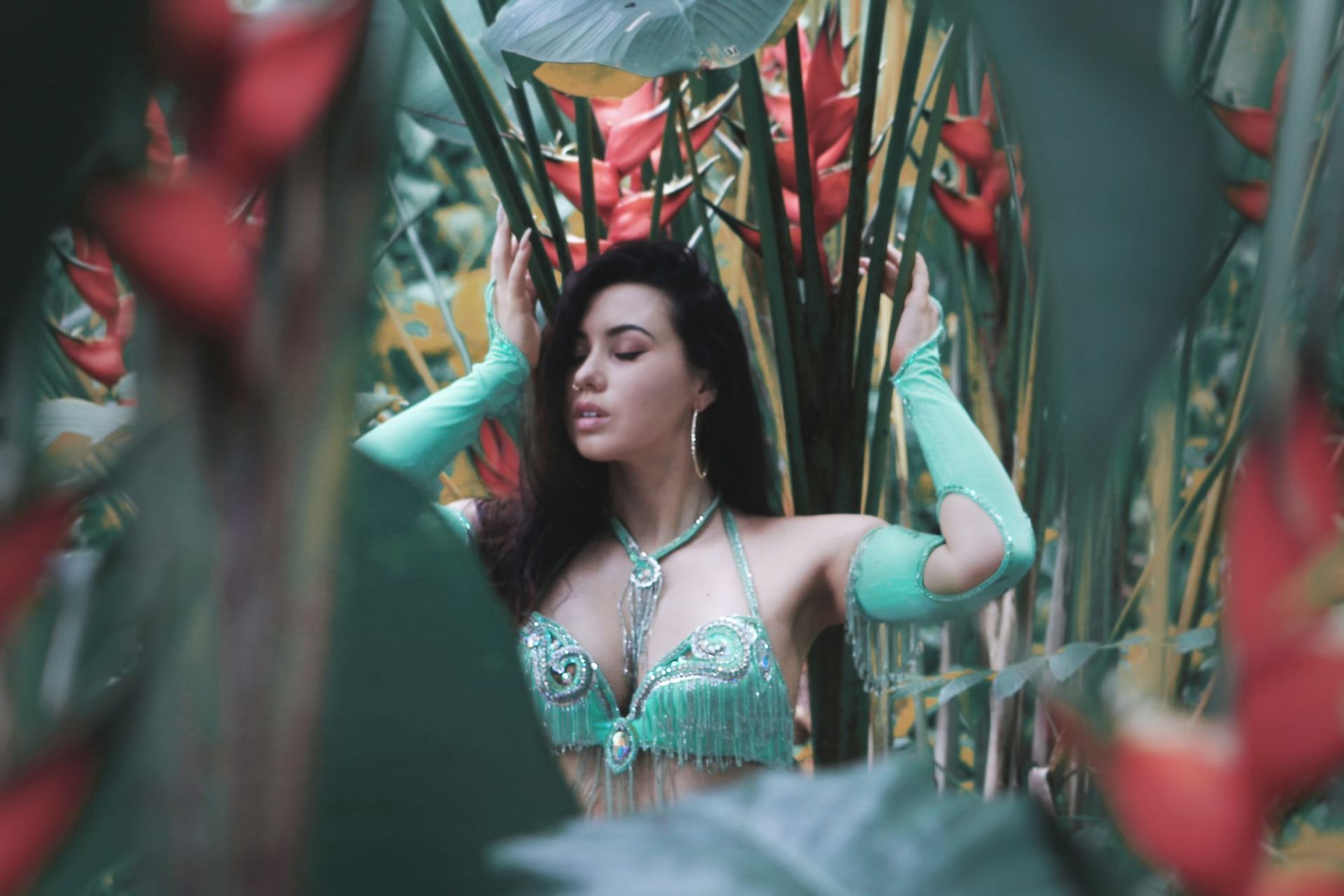 Belly dancer Azeeria Azizah in a green top and green gloves is standing in a jungle in Puerto Rico surrounded by flowers. 