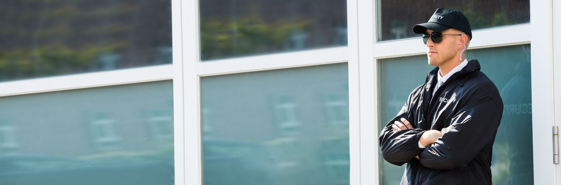 A security guard is standing outside a large windowed building with arms crossed