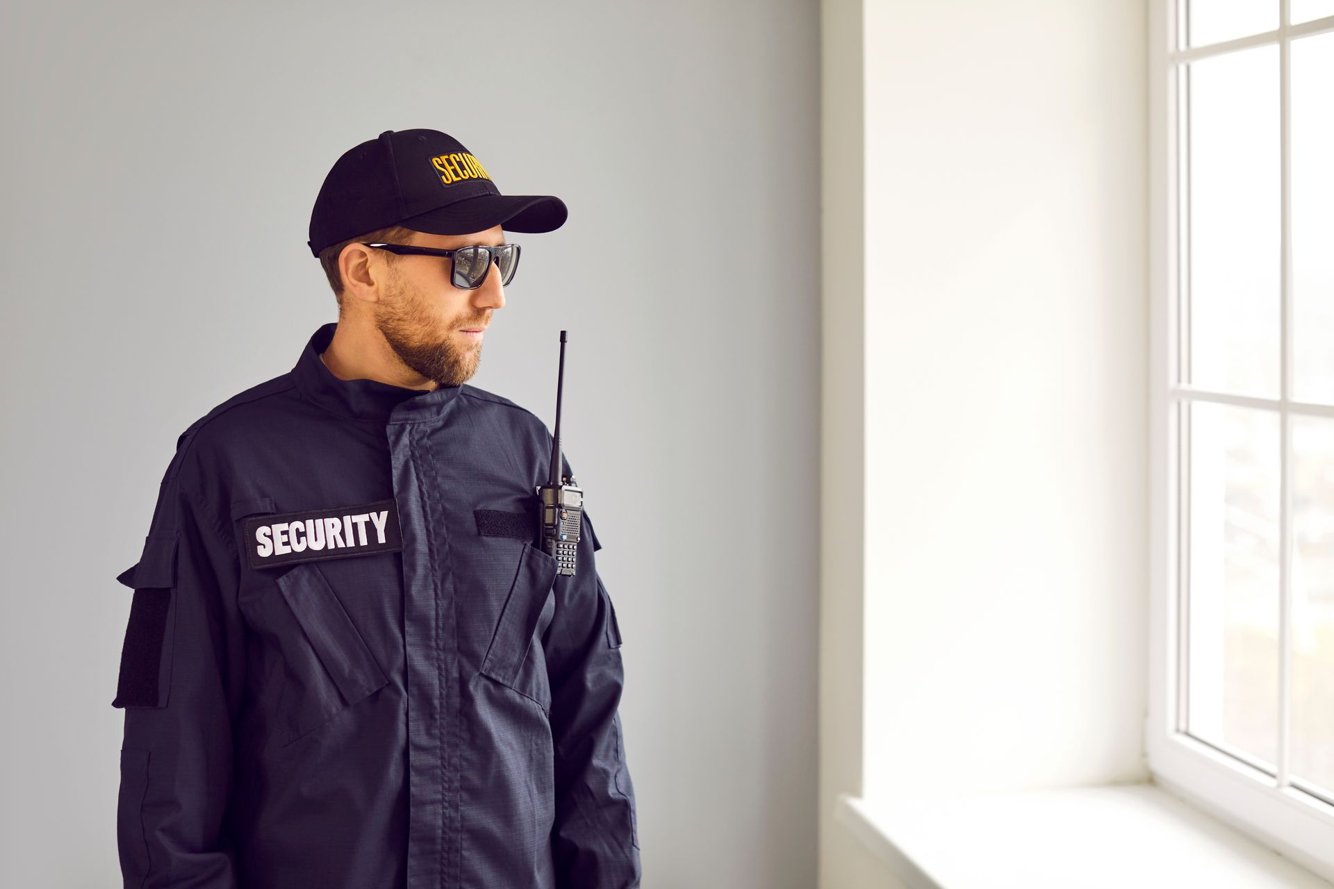 A security officer is standing by a window wearing a uniform and a radio.