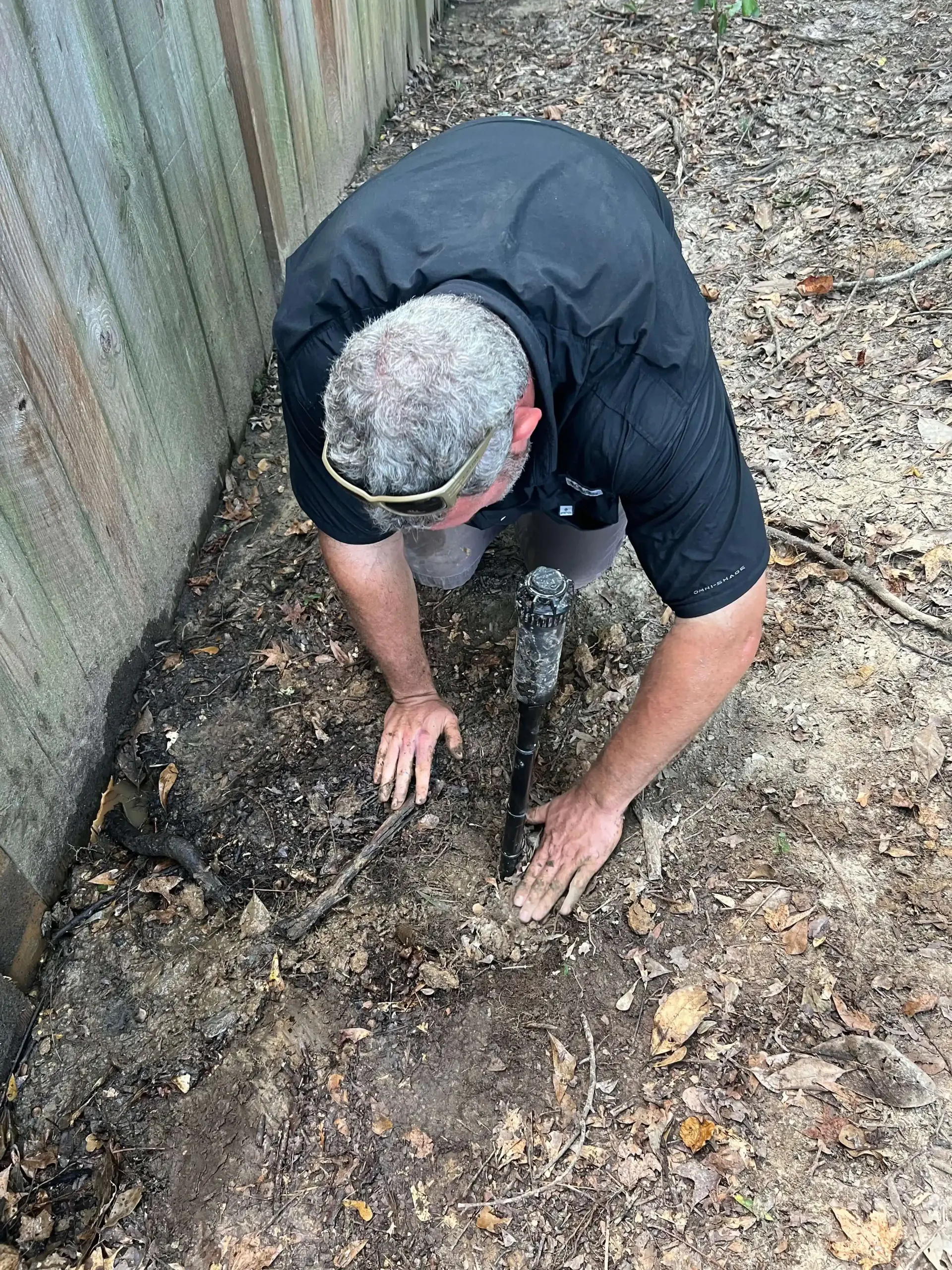 Man kneels, examining soil near a fence. Graying hair, black shirt, and sunglasses. Dirt ground.
