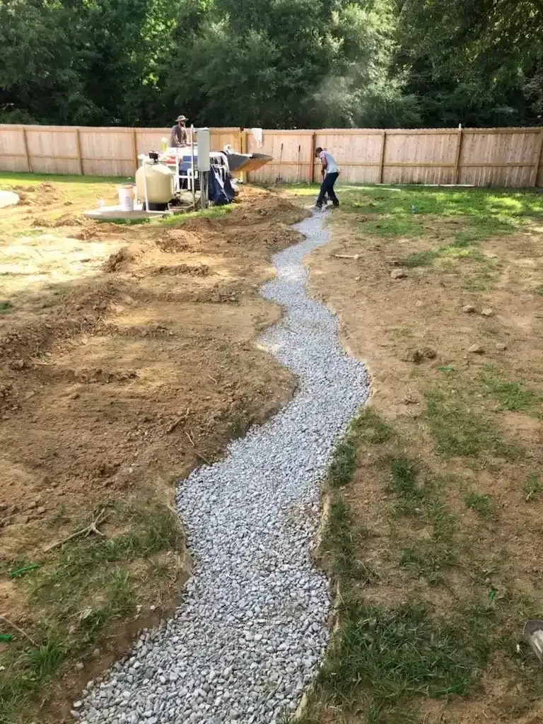 A gravel-filled trench being constructed in a backyard by workers near a fence and pool equipment.