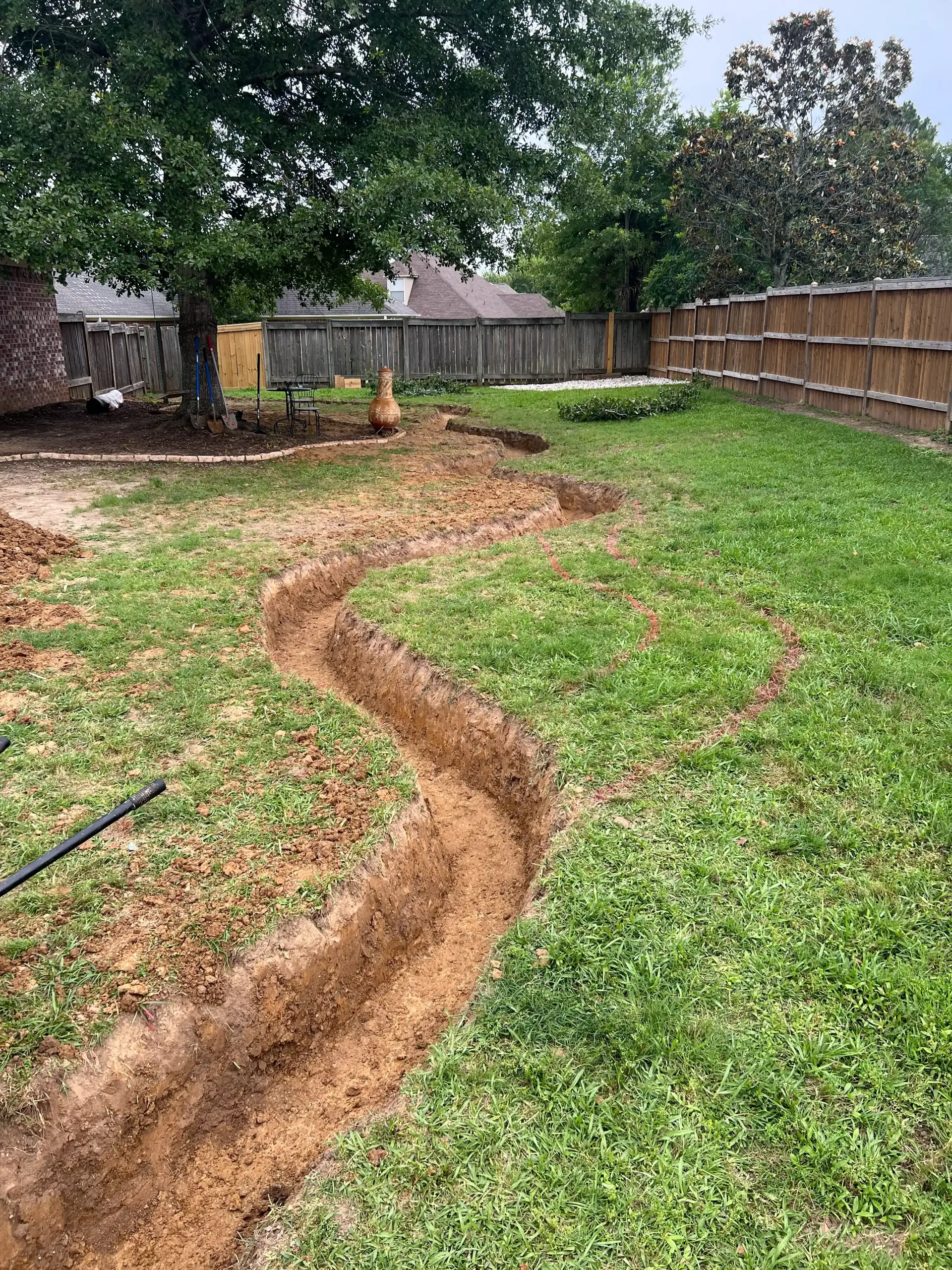 A winding trench dug in a grassy backyard, likely for laying pipes or cables, with a wooden fence in the background.