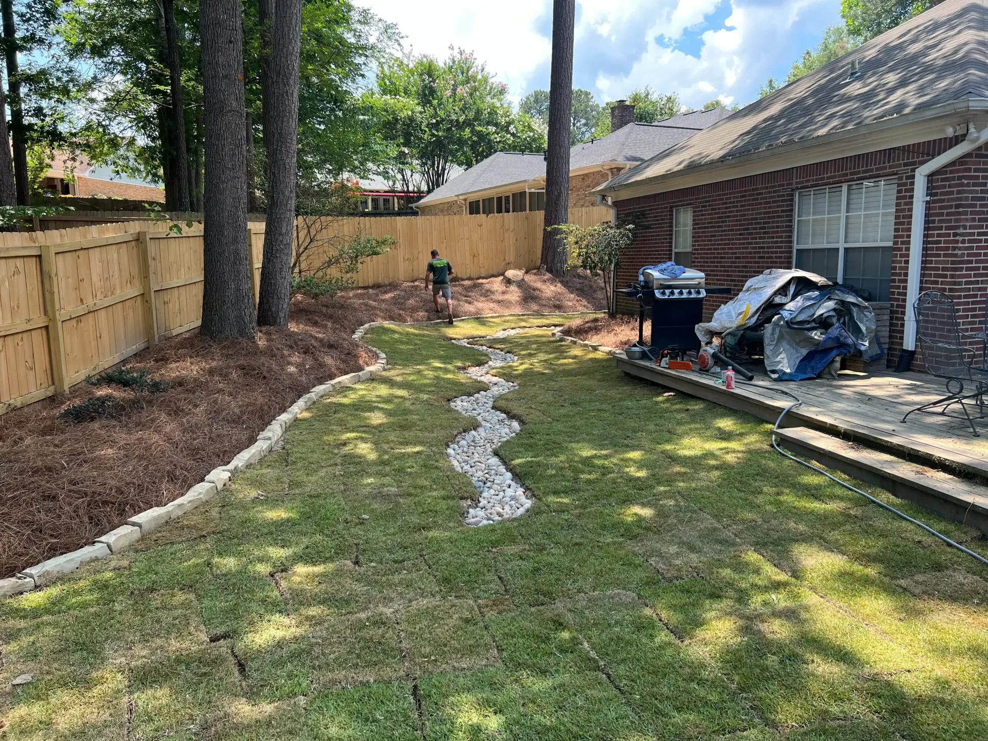 Backyard with a brick house, wooden fence, grass, gravel path, and grill.
