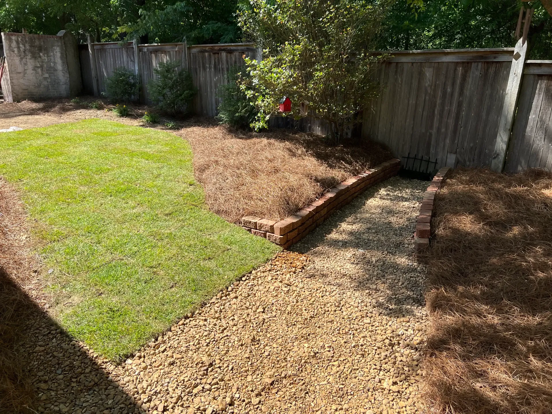 Backyard with gravel path, grass, and a mulch bed, bordered by a brick curb and wooden fence.