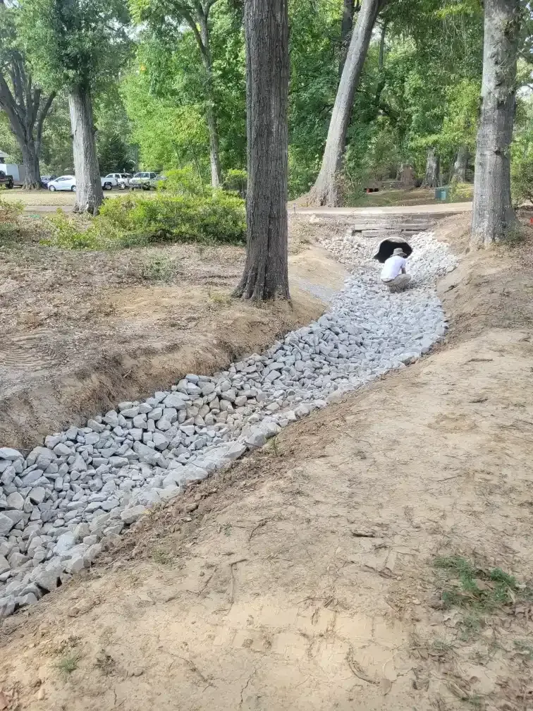 Gravel-lined ditch in a park, trees surrounding, person looking down.