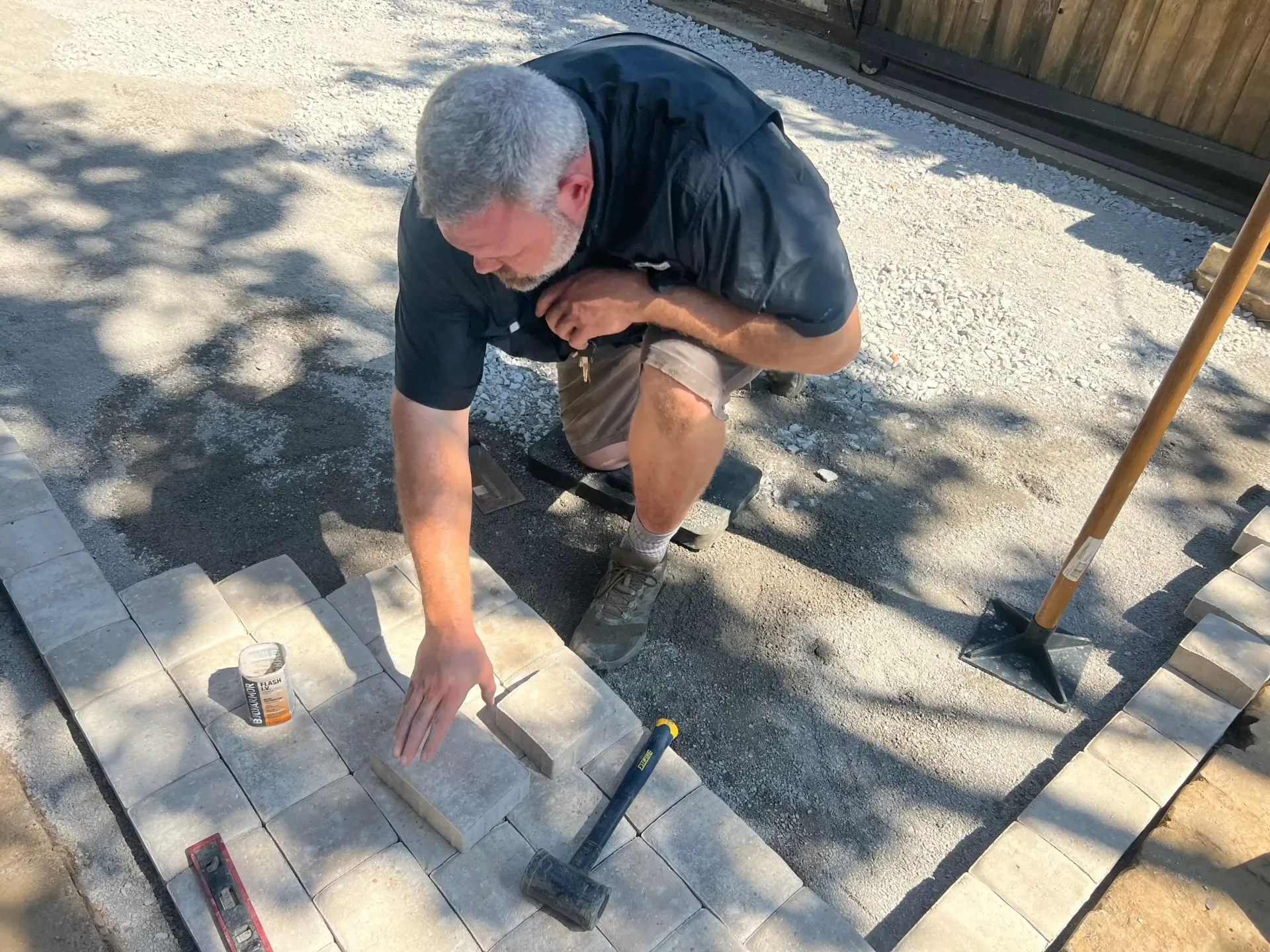 Man kneeling, laying pavers on gravel base; hammer and level visible. Outdoors.