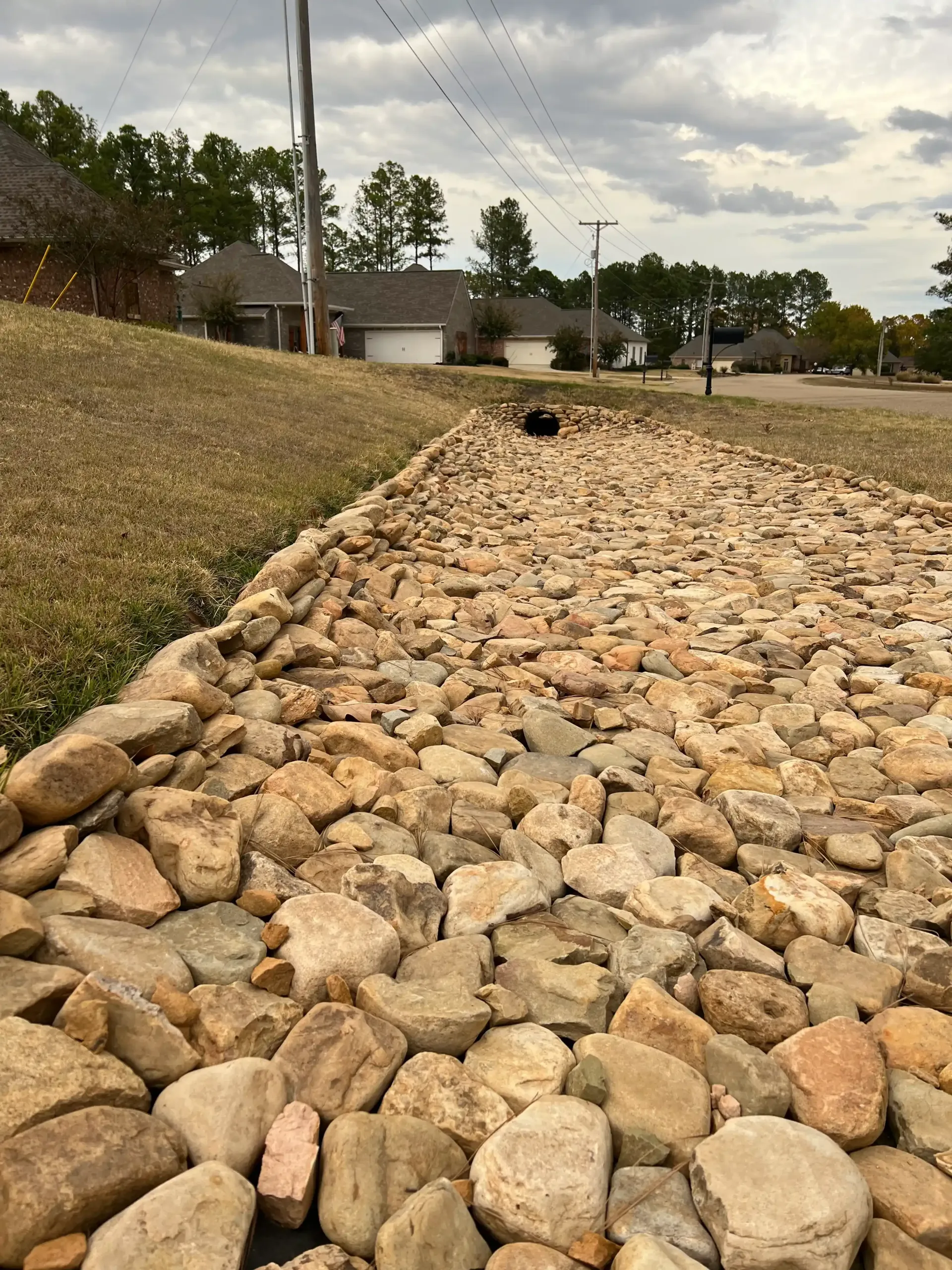 Rock-lined drainage ditch along a grassy slope in a residential area. Overcast sky.