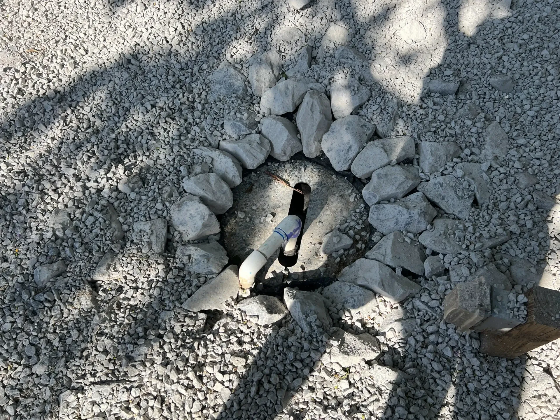 Black pipe emerging from ground surrounded by a circle of rocks set in gravel.