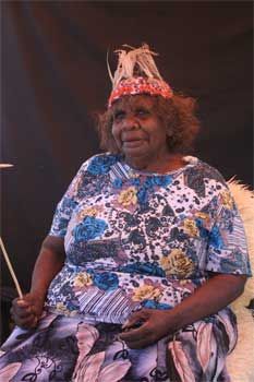 An elderly woman wearing a headband and a floral dress is sitting on a chair.