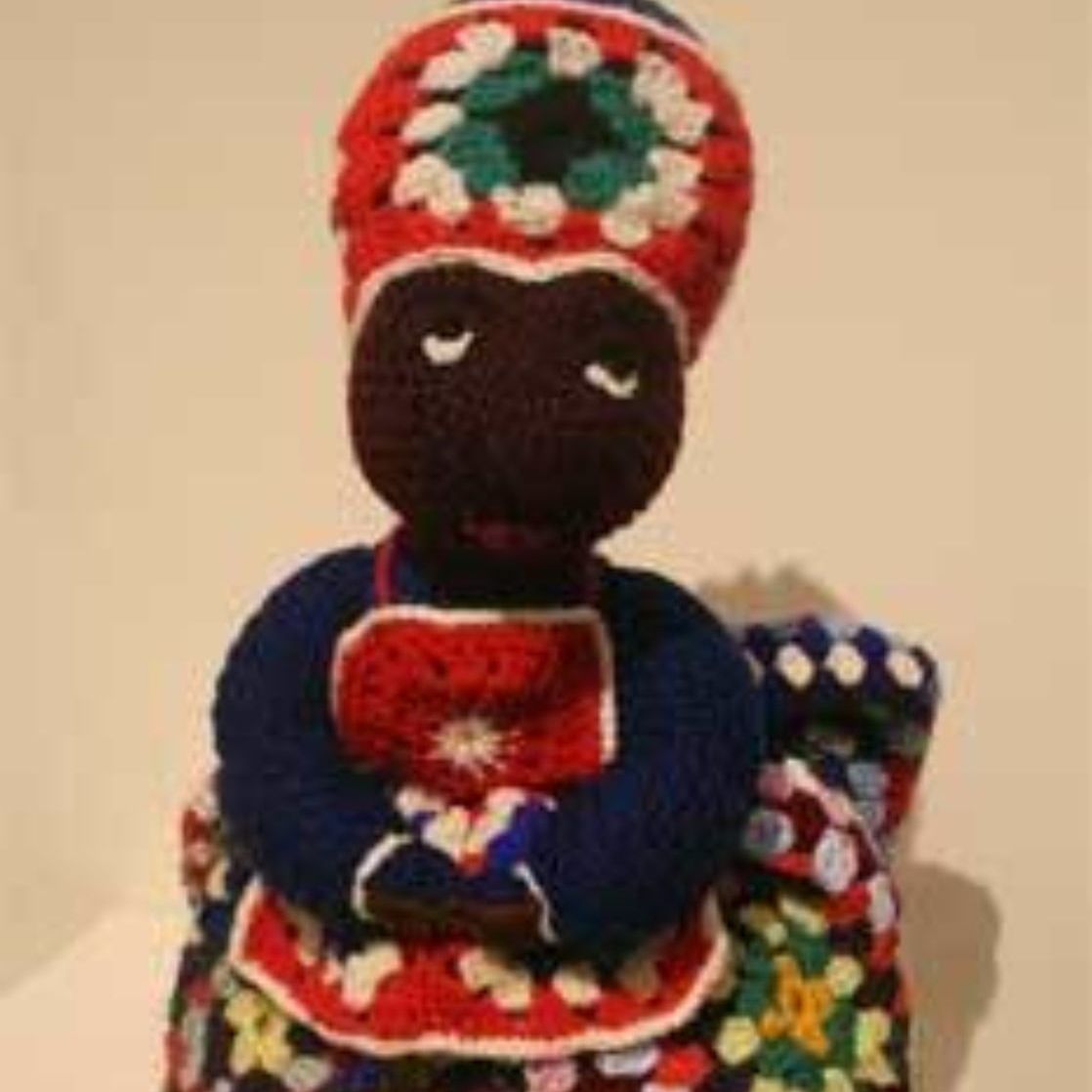 A crocheted doll is sitting on a blanket with its eyes closed.