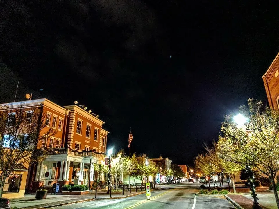 Nighttime street scene with illuminated buildings, trees, and streetlights under a dark sky.