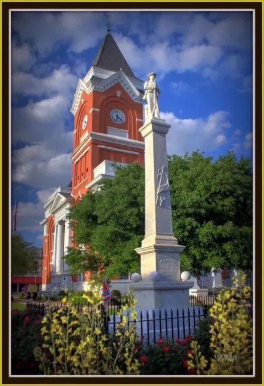 Red brick courthouse with clock tower and monument, flowers in foreground, blue sky.