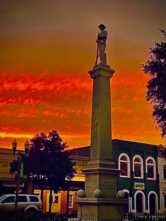 Monument of a soldier holding a rifle at sunset. Orange sky, green building, white car, and trees in the background.