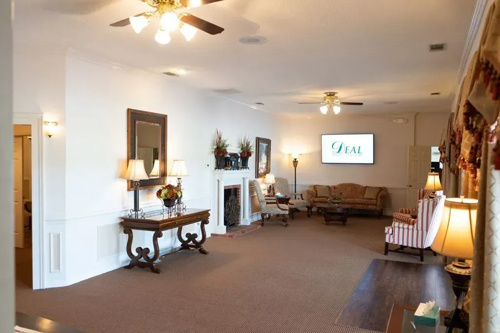 Spacious room, likely a funeral home, with seating, a fireplace, and a TV; neutral colors, carpeted floor.