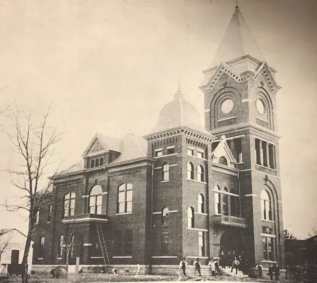 Brick courthouse with tower and steeple. Group of people on steps; tree on left.