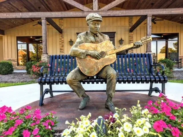 Statue of a man playing guitar seated on a bench, in front of a building, surrounded by flowers.