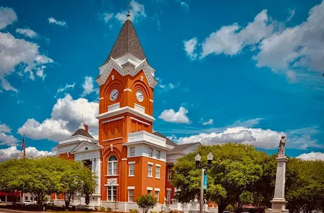 Red brick courthouse with clock tower against a blue sky, flanked by trees and a monument.