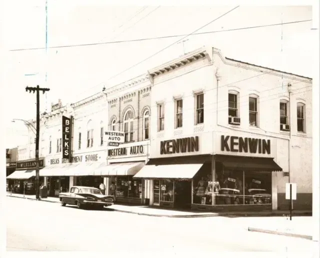 Black and white photo of a downtown street scene in front of buildings, with a vintage car parked on the street. 