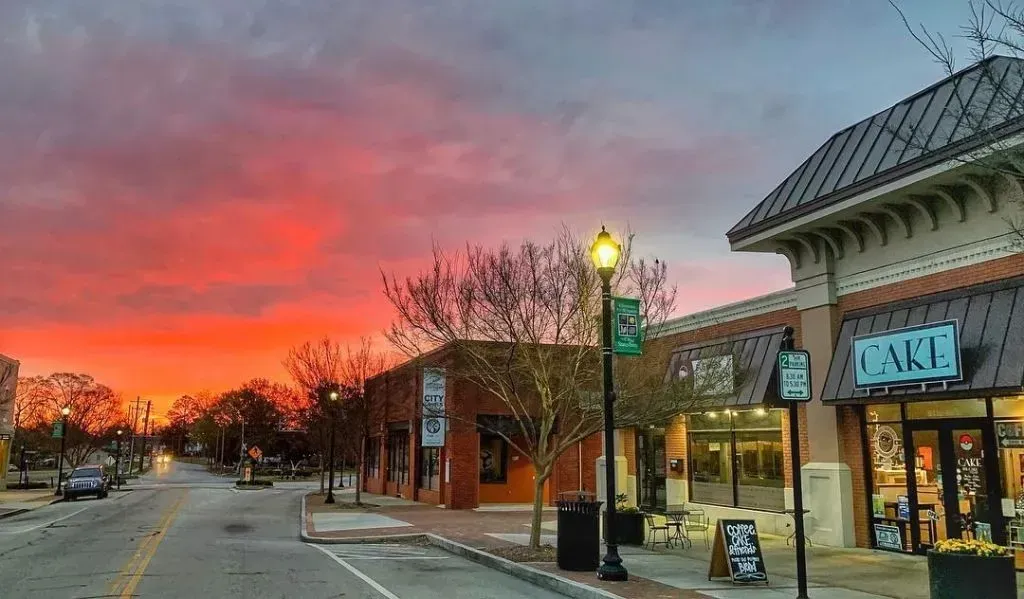 Street scene at sunset with buildings, trees, and glowing red sky. Storefronts line the street.