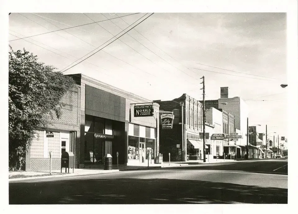 Street of shops with awnings, power lines overhead, and a tall building in the background; a sunny day.