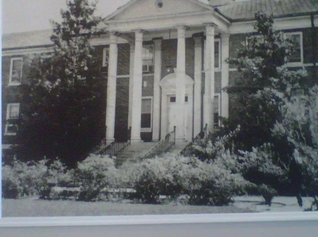 Black and white photo of a two-story building with large white columns and a small front yard with bushes.