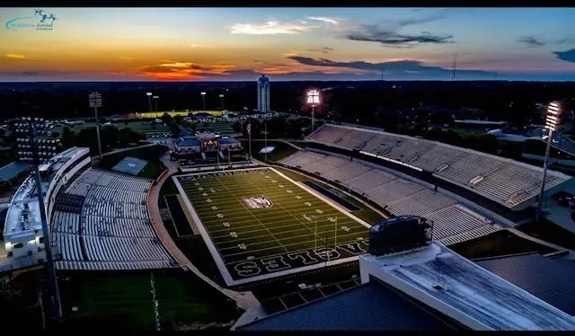 Aerial view of a football stadium at sunset with lit field and bleachers.