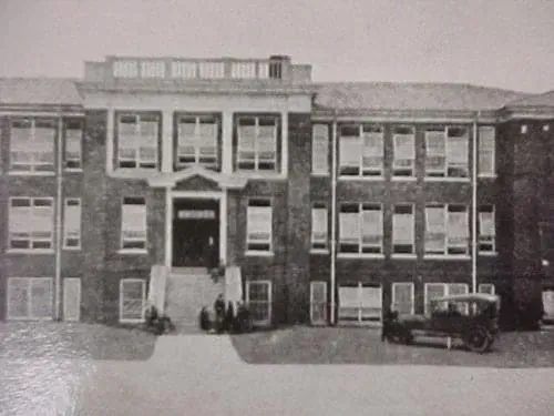 Two-story brick schoolhouse with multiple windows, central entrance, and a vintage car parked out front.
