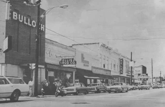 Street scene in black and white with vintage cars parked along the curb. Signs for shops are visible, including 