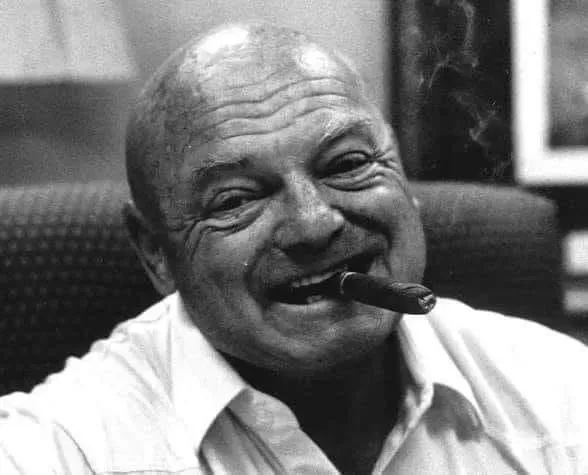 Bald man with cigar smiling, smoke rising, seated.