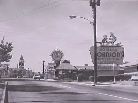 Black and white photo of a street with a Howard Johnson's restaurant and sign, a church, and cars.