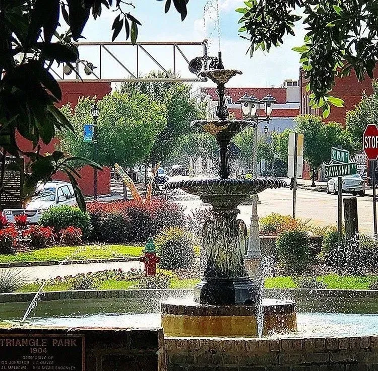 Fountain in Triangle Park, brick base, water spraying, with street and buildings in the background.