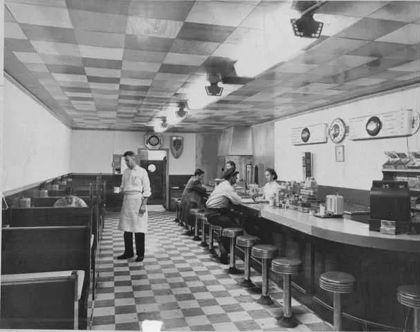 Interior view of a diner with checkered floor and ceiling. A counter with stools runs alongside a line of booths. A server stands near the booths.