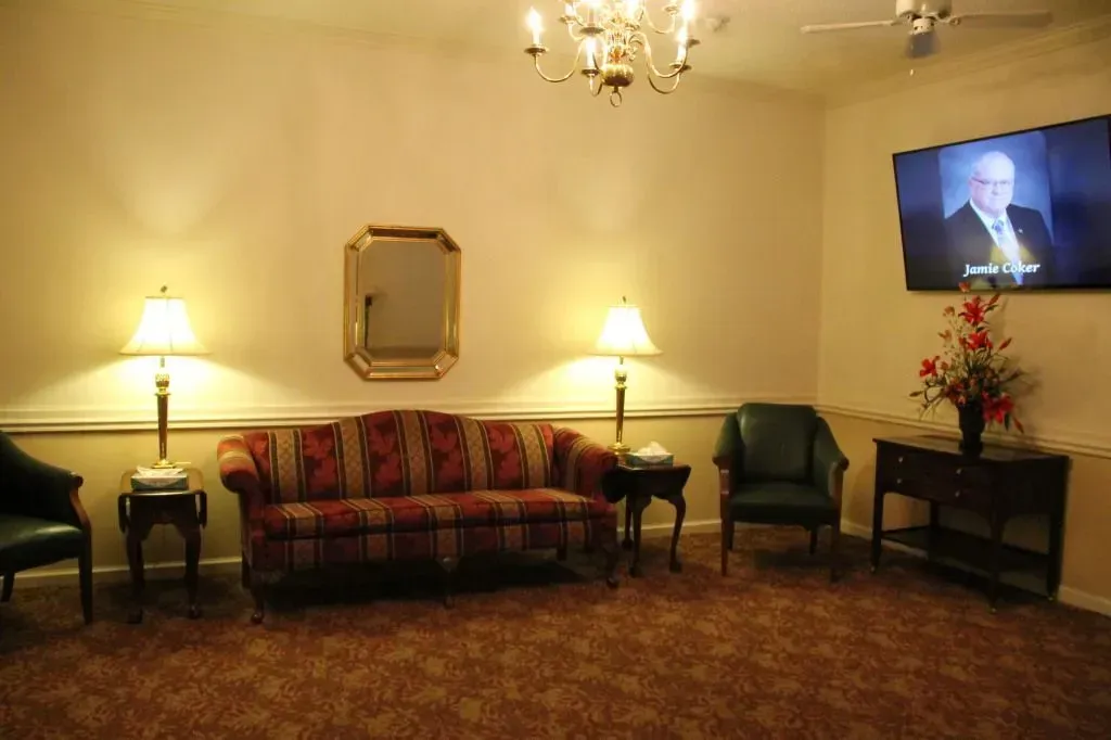Funeral home waiting area with sofa, chairs, lamps, mirror, TV, floral arrangement.