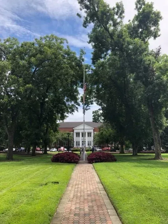 Brick path leading to a white building with columns, flanked by trees and red bushes, set on a green lawn.