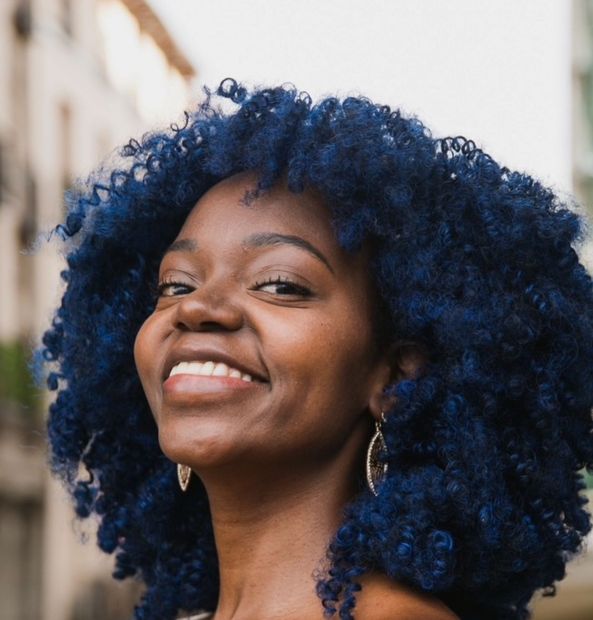 Smiling Black woman with blue afro, in an outdoor setting, wearing earrings.