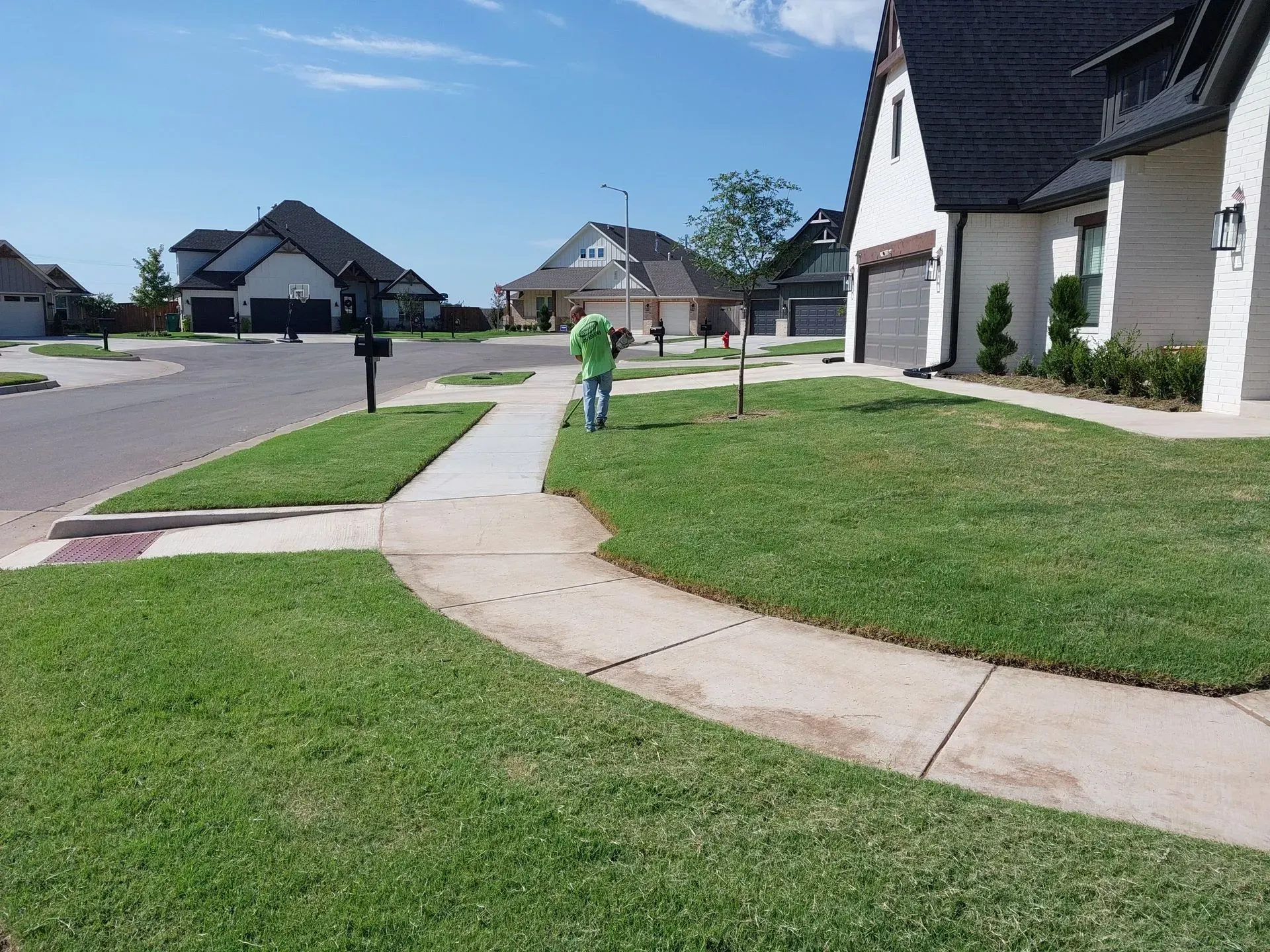 Person mowing a lush green lawn next to a sidewalk in a suburban neighborhood on a sunny day.