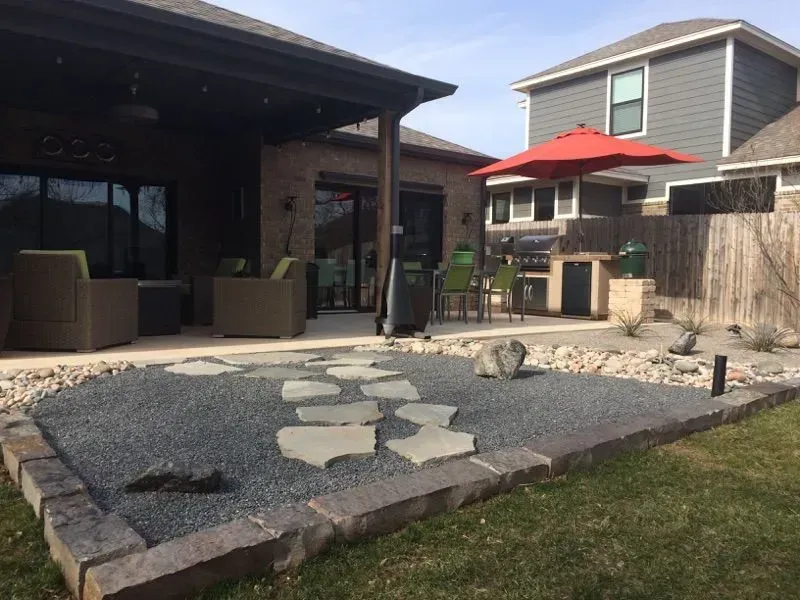 Patio with stone path, gravel, outdoor kitchen, seating under a covered area, and red umbrella.