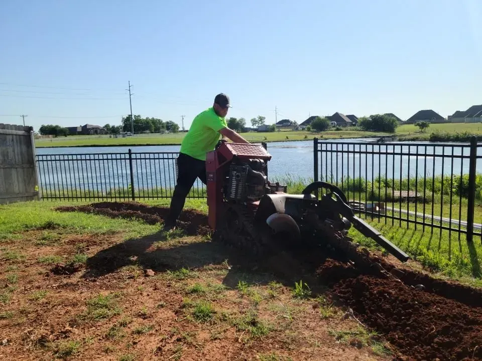 Man operating a trencher along a fence, near a pond. Bright green shirt, sunny day.