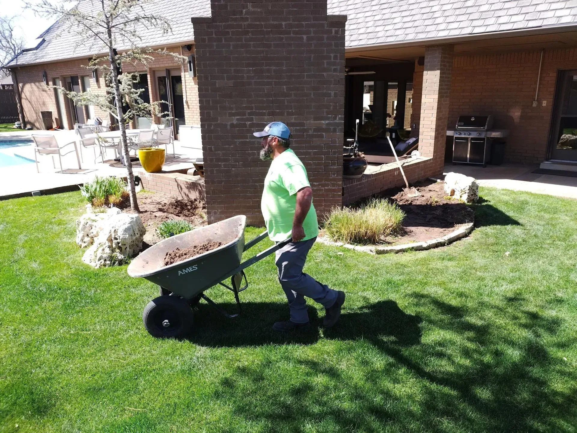 Man in green shirt pushes a wheelbarrow full of debris across a green lawn near a brick structure and pool.
