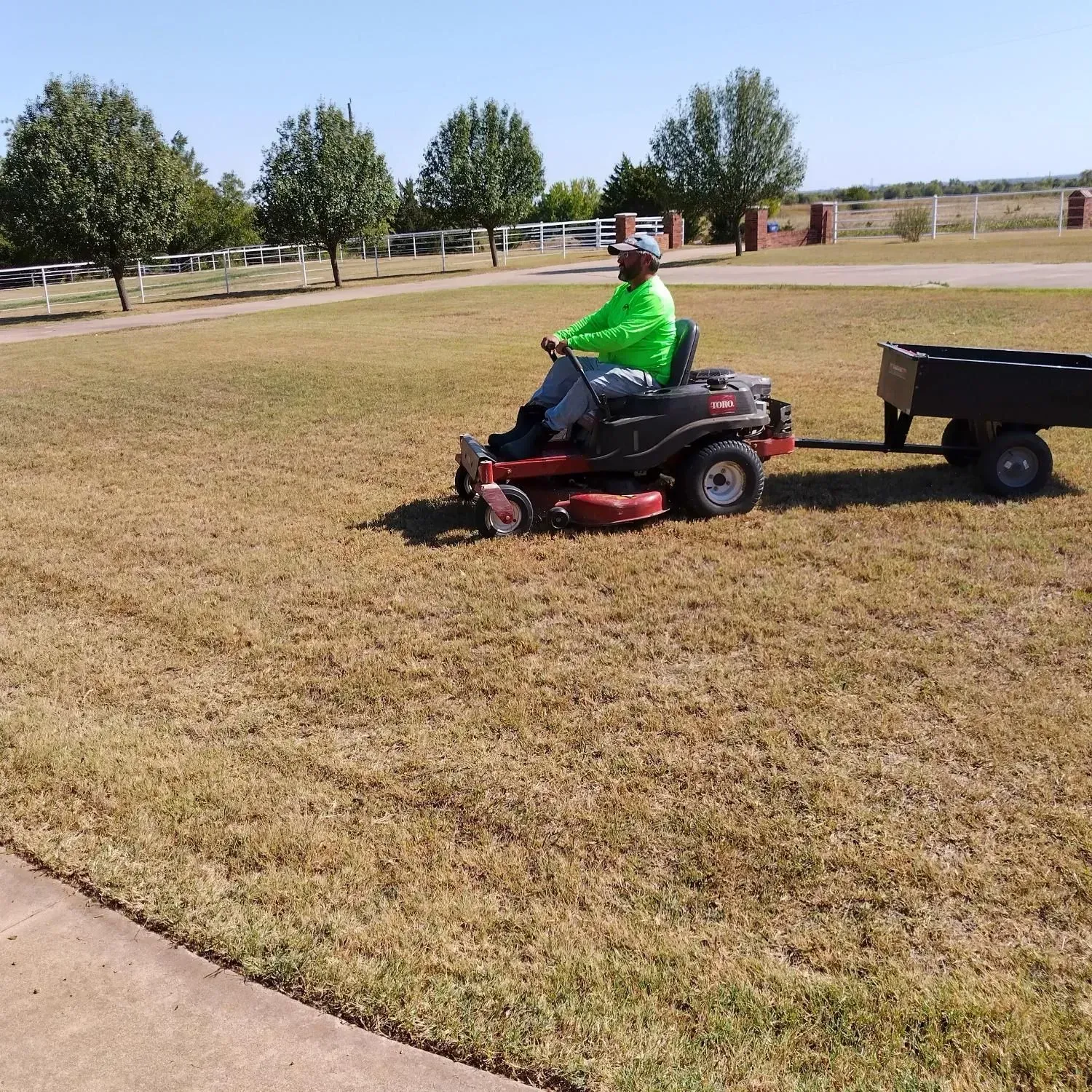 Person mowing lawn with riding mower and attached trailer on a sunny day.