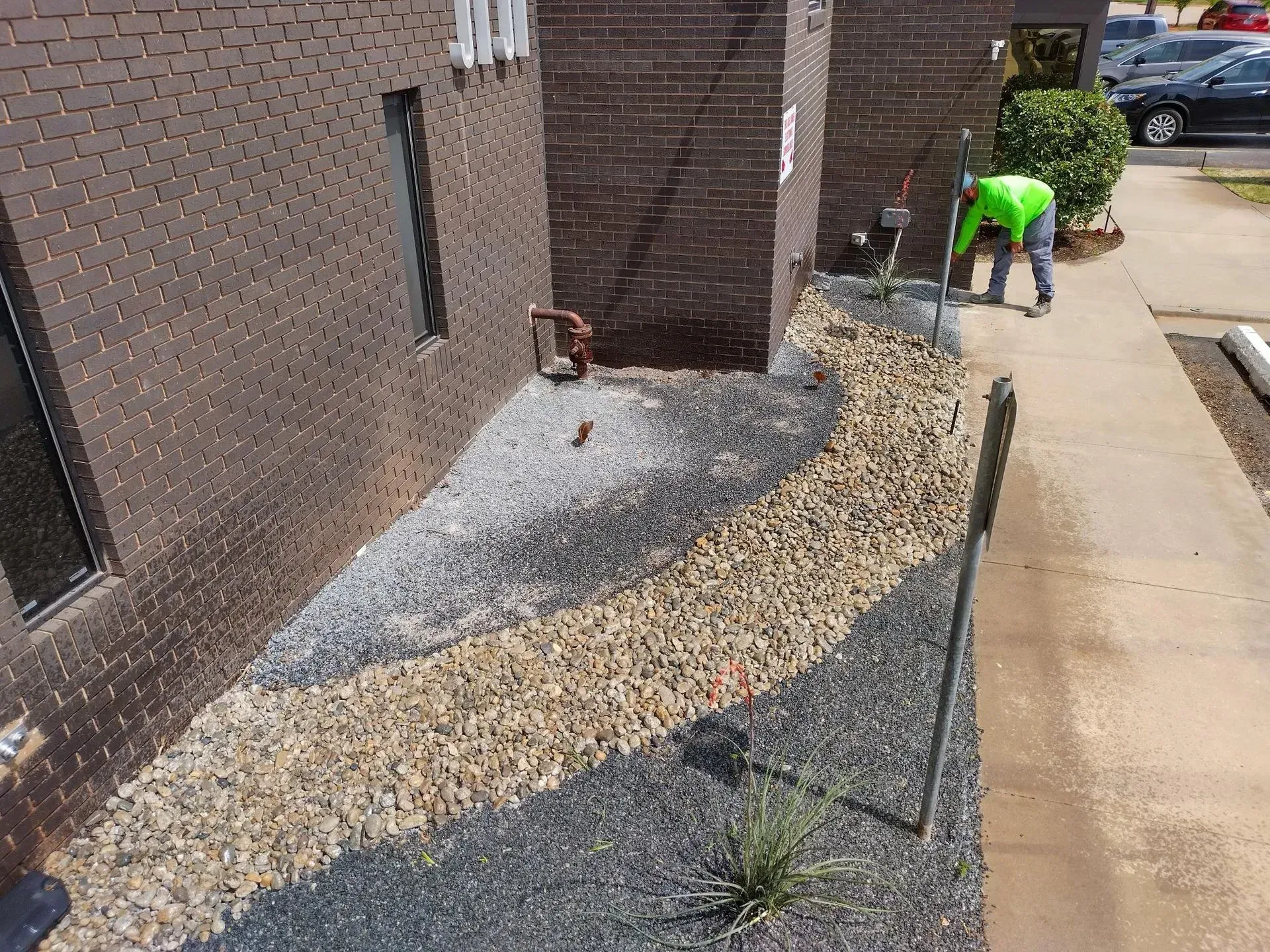 Person in neon green shirt working on a gravel walkway beside a brick building.
