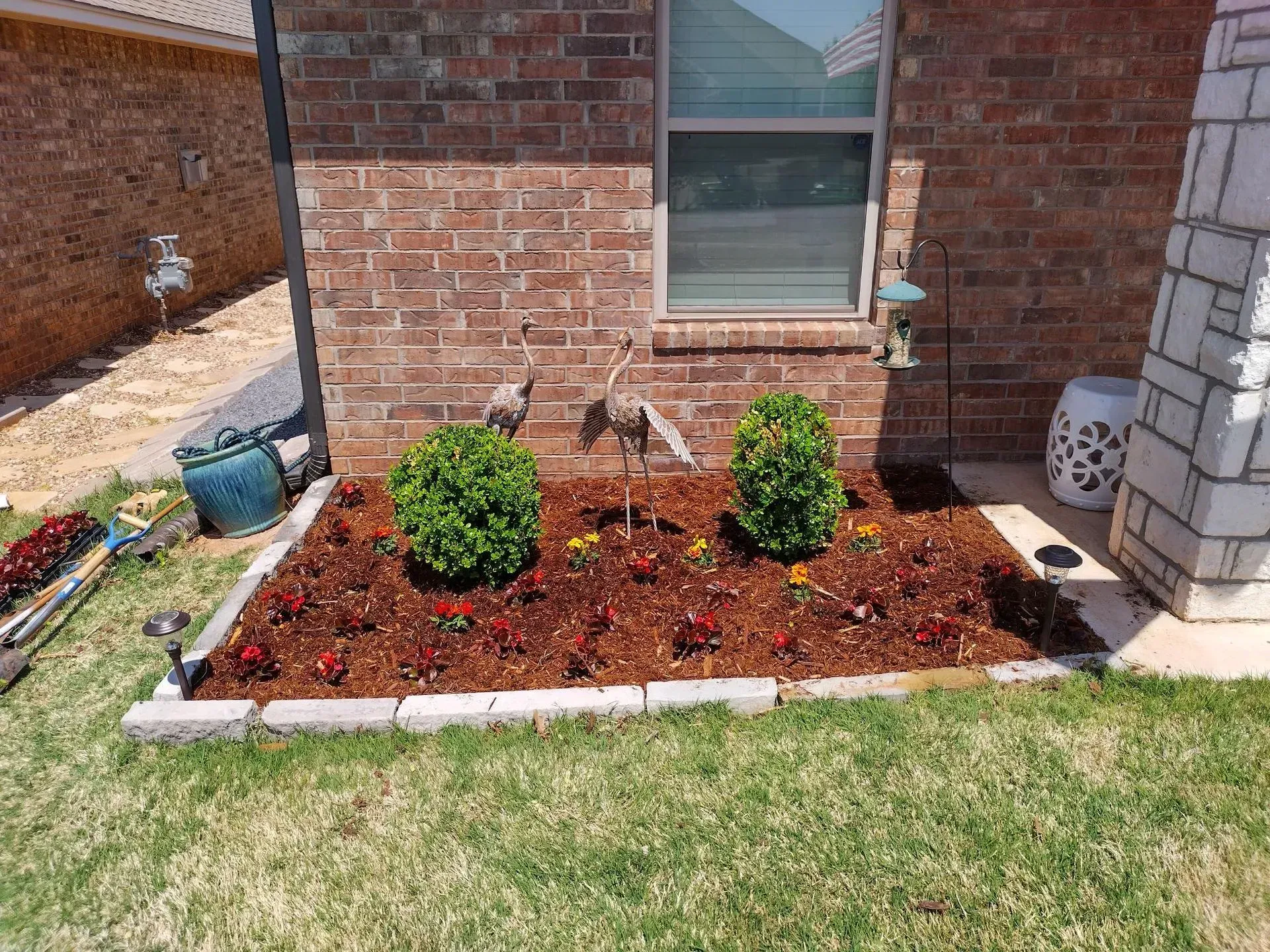 Flower bed with red mulch, green bushes, and small flowers in front of a brick building.