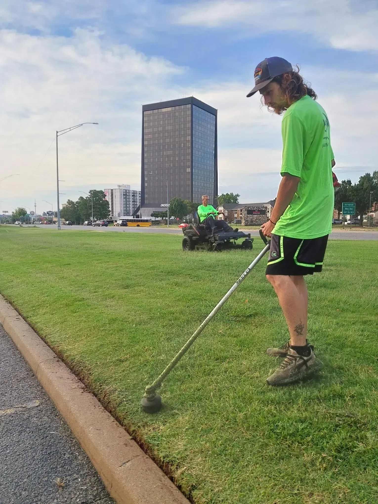 Person using a string trimmer to edge a lawn next to a road, tall building in the background.