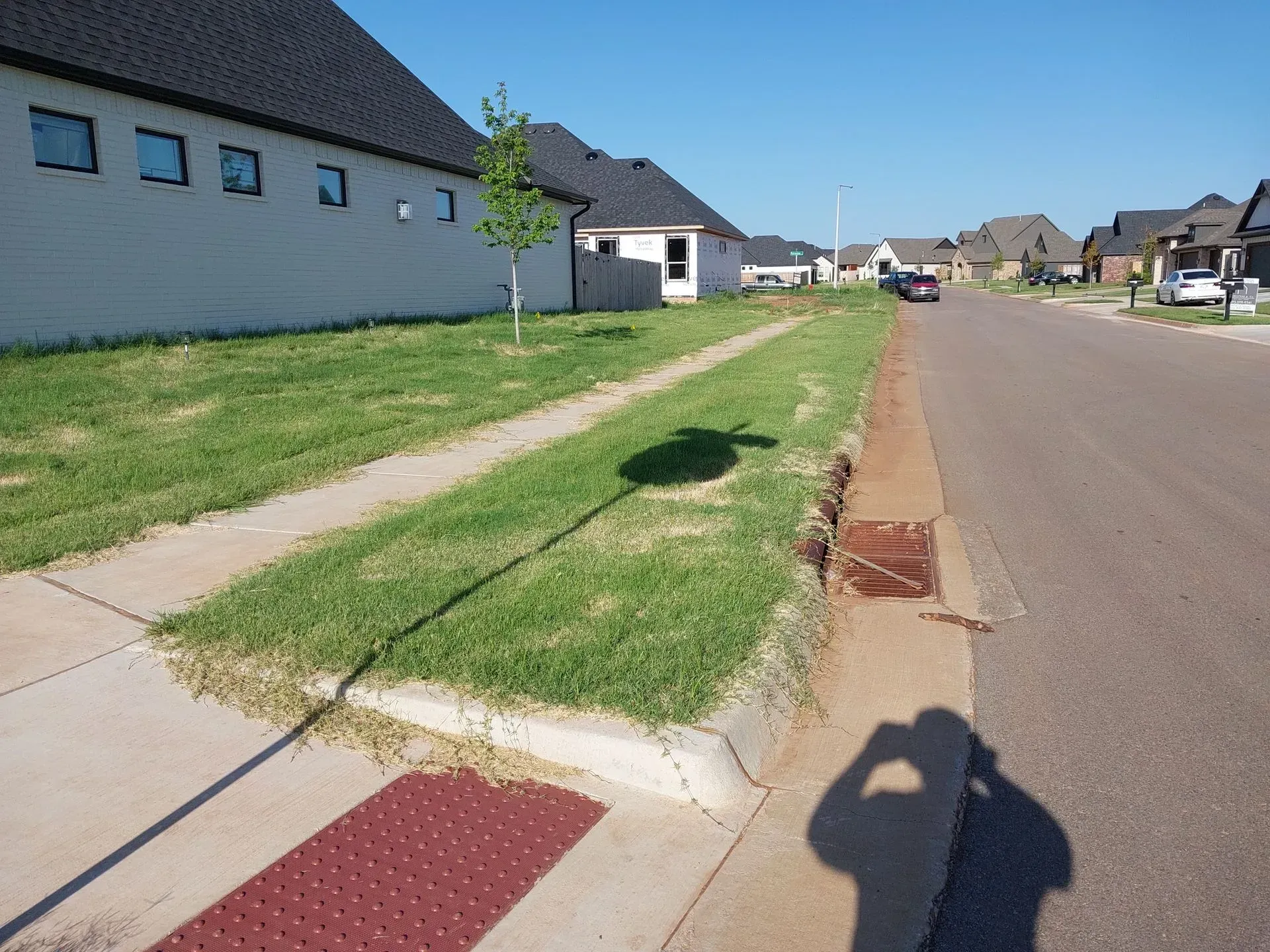 Sidewalk and grass verge along a street with houses; sunny day. Red tactile paving at crosswalk.